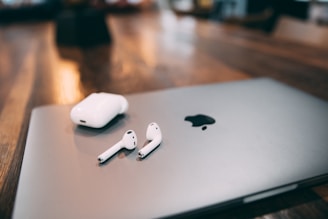 A set of wireless earphones along with their charging case rests on a sleek, silver laptop with an apple logo. The wooden surface background adds a warm contrast to the modern, metallic devices.