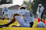 A football player in a white jersey and shorts sits on the grass with a helmet beside him, appearing to rest or take a break. Several other players and red cones are visible in the background, suggesting a practice session on a sunny day.