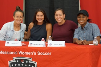 Four people sit at a table covered with a red cloth that reads 'Professional Women's Soccer.' They are smiling and appear to be at an event or press conference. Name cards are visible in front of each attendee.