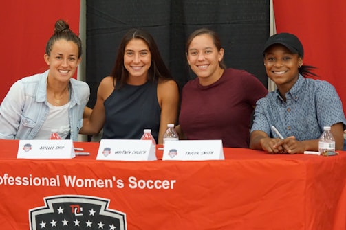 Four people sit at a table covered with a red cloth that reads 'Professional Women's Soccer.' They are smiling and appear to be at an event or press conference. Name cards are visible in front of each attendee.