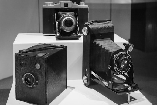 Three vintage cameras are displayed on white pedestals, featuring a box camera, a folding camera, and a compact camera. The image is in black and white, emphasizing the retro aesthetic of the cameras.