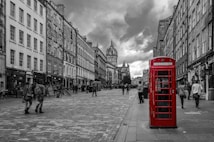 A vibrant red telephone booth stands prominently on a cobblestone street, surrounded by historical buildings with detailed architecture. The scene is set against a dramatic, cloudy sky, with people casually walking along the street.