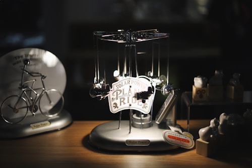 A solar-powered model of a plane ride is placed on a wooden surface. It has a sign on it indicating 'SOLAR POWER PLANE RIDE'. Next to it on the left, there is a solar-powered model of a bicycle and on the right, there are small figurines and objects. The overall scene is dimly lit, with light focused on the central plane ride model.