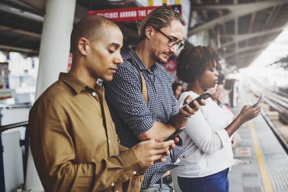 two men and one woman standing on station while holding mobile phones during daytime