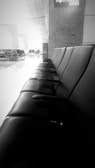 A row of empty black leather chairs positioned in a straight line within a waiting area or terminal. The space is modern, with clean lines and a high ceiling. There are more chairs and seating areas visible in the background, alongside large structural columns and windows.