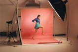 A young woman is posing confidently in a photography studio. She is wearing a dark blue top, a turquoise skirt, and red high-heeled shoes, which match the pink background. The studio setup includes large softbox lights and a reflector, giving a professional and vibrant atmosphere.