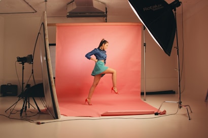A young woman is posing confidently in a photography studio. She is wearing a dark blue top, a turquoise skirt, and red high-heeled shoes, which match the pink background. The studio setup includes large softbox lights and a reflector, giving a professional and vibrant atmosphere.