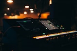 A dimly lit environment with a focus on an illuminated sound mixing console. The console's lights and screens stand out against the dark background, hinting at an audio or concert setup.