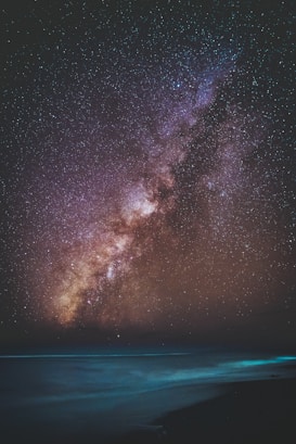 A vibrant and richly colored night sky filled with countless stars, showcasing the Milky Way galaxy in a stunning display of purples, blues, and whites. Below the celestial scene is a dark, tranquil ocean with a faint blue glow on the horizon, indicating a subtle luminescence.