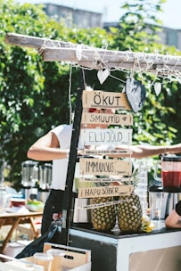 A smoothie stand outdoors featuring handwritten signs with smoothie names in an unidentified language. Pineapples are displayed, and a person is preparing drinks using a blender. The area is surrounded by greenery and a sunny atmosphere is evident.