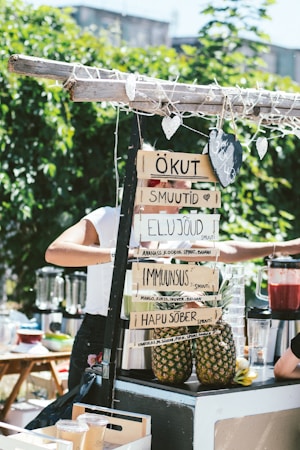 A smoothie stand outdoors featuring handwritten signs with smoothie names in an unidentified language. Pineapples are displayed, and a person is preparing drinks using a blender. The area is surrounded by greenery and a sunny atmosphere is evident.
