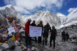 A group of people is gathered at Everest Base Camp in front of a mountainous backdrop. They hold a banner with colorful prayer flags piled on the left. The sky is partly cloudy, adding to the rugged mountainous scenery.