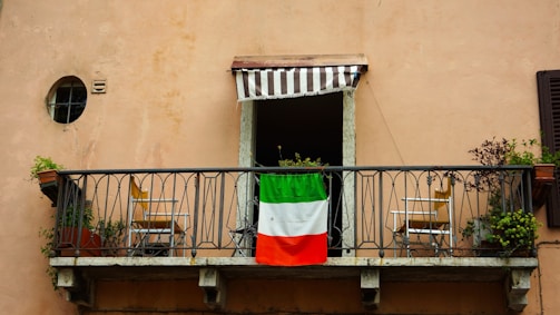 A small balcony features an iron railing with an Italian flag draped over it. Two wooden chairs are placed symmetrically on either side of the balcony, each accompanied by potted plants. Above the balcony is a small window with a striped awning.