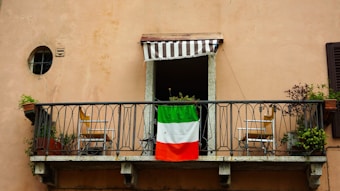 A small balcony features an iron railing with an Italian flag draped over it. Two wooden chairs are placed symmetrically on either side of the balcony, each accompanied by potted plants. Above the balcony is a small window with a striped awning.