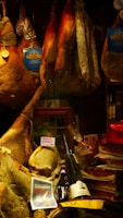 A display of various cured meats, including whole legs of ham, sausages, and salami, hanging alongside packaged products. Below, there are bottles of wine and a ribbon-wrapped item. Labels like 'Petit Marcel' and 'Prosciutto di Norcia' are visible, suggesting a focus on quality and origin.