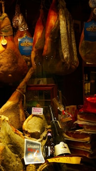 A display of various cured meats, including whole legs of ham, sausages, and salami, hanging alongside packaged products. Below, there are bottles of wine and a ribbon-wrapped item. Labels like 'Petit Marcel' and 'Prosciutto di Norcia' are visible, suggesting a focus on quality and origin.