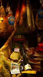 A display of various cured meats, including whole legs of ham, sausages, and salami, hanging alongside packaged products. Below, there are bottles of wine and a ribbon-wrapped item. Labels like 'Petit Marcel' and 'Prosciutto di Norcia' are visible, suggesting a focus on quality and origin.