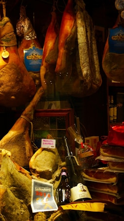 A display of various cured meats, including whole legs of ham, sausages, and salami, hanging alongside packaged products. Below, there are bottles of wine and a ribbon-wrapped item. Labels like 'Petit Marcel' and 'Prosciutto di Norcia' are visible, suggesting a focus on quality and origin.