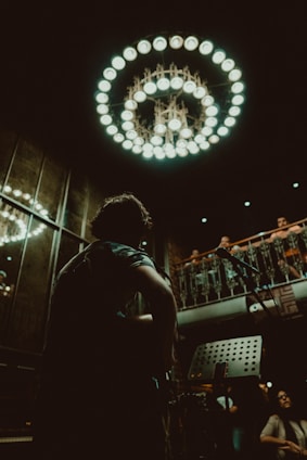 A musician with a guitar is in the foreground, silhouetted against a dimly lit interior adorned with an ornate chandelier. The setting appears to be a small performance venue with an audience on an upper balcony, giving a sense of intimacy and focus on live music.
