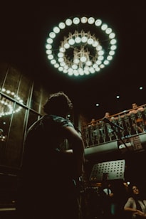 A musician with a guitar is in the foreground, silhouetted against a dimly lit interior adorned with an ornate chandelier. The setting appears to be a small performance venue with an audience on an upper balcony, giving a sense of intimacy and focus on live music.