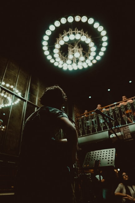 A musician with a guitar is in the foreground, silhouetted against a dimly lit interior adorned with an ornate chandelier. The setting appears to be a small performance venue with an audience on an upper balcony, giving a sense of intimacy and focus on live music.