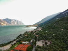 A picturesque landscape featuring a large body of water bordered by mountainous terrain. A village with red rooftops is nestled along the water's edge, surrounded by lush greenery and trees. There are tennis courts visible in the foreground, and the view stretches towards the horizon with dramatic mountains in the backdrop.
