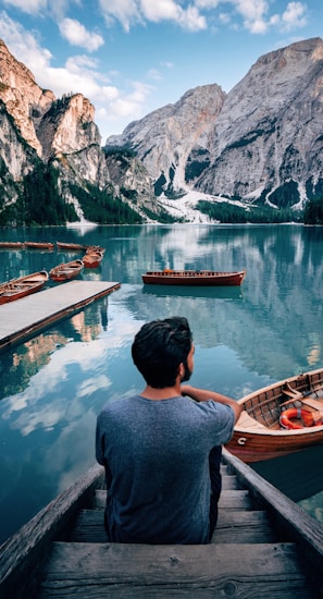 man sitting on brown wooden stairs staring out lake near mountain during daytime