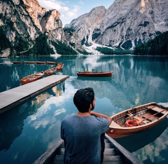 man sitting on brown wooden stairs staring out lake near mountain during daytime