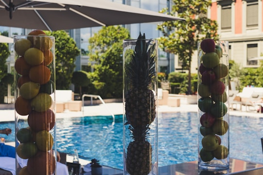 Tall glass vases filled with assorted fruits including apples, oranges, and a pineapple are displayed on a countertop beside a clear, blue swimming pool. Large umbrellas provide shade, and lush greenery surrounds the pool area, giving a sense of luxury and relaxation.