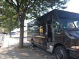 A UPS delivery truck parked on the side of a street under a tree. The truck is brown with the UPS logo prominently displayed on the side. The street is lined with a sidewalk and there are shaded areas due to the tree's foliage.