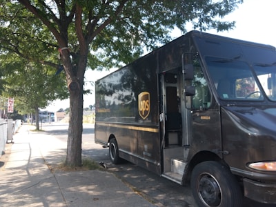 A UPS delivery truck parked on the side of a street under a tree. The truck is brown with the UPS logo prominently displayed on the side. The street is lined with a sidewalk and there are shaded areas due to the tree's foliage.