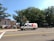 A FedEx Express delivery van is parked on the side of a street. The foreground features a road with yellow and white lane markings. In the background, there are several tall leafy trees and a row of commercial buildings, some with red brick facades. A small park or public square is visible, with people walking and a few tents or stalls set up.