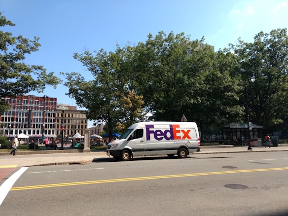 A FedEx Express delivery van is parked on the side of a street. The foreground features a road with yellow and white lane markings. In the background, there are several tall leafy trees and a row of commercial buildings, some with red brick facades. A small park or public square is visible, with people walking and a few tents or stalls set up.