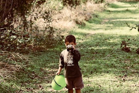 A young child stands on a grassy path in a wooded area, holding a pair of binoculars to their eyes and a bright green bucket in the other hand. Surrounding the path are dense trees and foliage, with the sunlight filtering through, creating a serene natural environment.