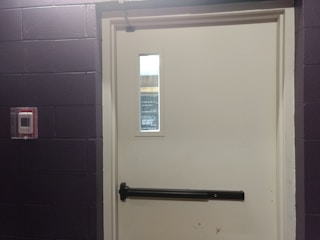 A closed white door set in a dark-colored brick wall, featuring a push bar and a window near the top. To the left of the door, there is a red fire alarm device mounted on the wall.