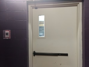 A closed white door set in a dark-colored brick wall, featuring a push bar and a window near the top. To the left of the door, there is a red fire alarm device mounted on the wall.