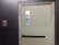 A closed white door set in a dark-colored brick wall, featuring a push bar and a window near the top. To the left of the door, there is a red fire alarm device mounted on the wall.