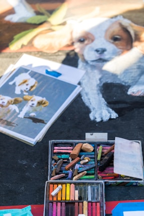 An assortment of colorful pastel sticks is arranged in a box, accompanied by a drawing in progress depicting a puppy. Beside the artwork, there are pages on a clipboard featuring images of puppies, likely serving as reference material.