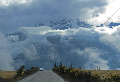 A winding road leads towards a majestic mountain partially shrouded in dense clouds. The mountainous landscape is framed by an expansive sky filled with dramatic cloud formations. A car travels down the road amid lush vegetation on either side, creating a sense of solitude and adventure.