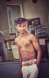 A young man with short, dark hair poses shirtless, leaning casually against a glass surface. He wears light-gray trousers and a brown belt, with earphones draped around his neck. Behind him, there is a blurred view of a modern interior with chairs and a counter displaying several bottles.