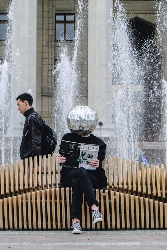 person sitting on wooden accordion bench while reading magazine at the back of water fountain in Almaty Kazakhstan