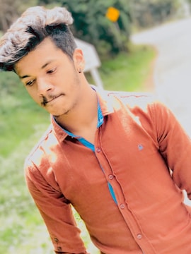 A young person with styled hair is wearing a rust-colored button-down shirt with a blue inner lining. They appear to be outdoors, with a blurred background of greenery and a road.