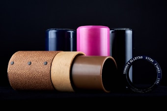 A collection of cylindrical objects in various materials and colors is arranged on a dark background. The objects include textured, metallic, and glossy surfaces in shades of brown, pink, and black. The arrangement is orderly, with the cylinders standing upright and one lying horizontally in the foreground.