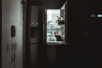 The scene depicts a dimly lit kitchen interior with the door of a refrigerator left open, casting a cool white glow. Inside the fridge, various items such as bottles and containers are visible. A wall-mounted telephone is seen to the left, contributing to the ambiance of the room.