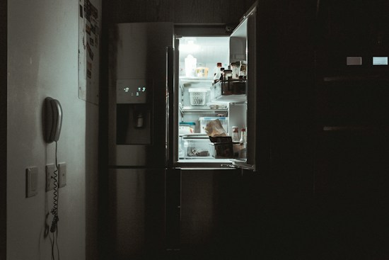 The scene depicts a dimly lit kitchen interior with the door of a refrigerator left open, casting a cool white glow. Inside the fridge, various items such as bottles and containers are visible. A wall-mounted telephone is seen to the left, contributing to the ambiance of the room.