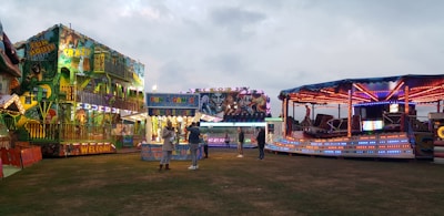 A lively amusement park scene features brightly lit rides and attractions. A fun house with vibrant lights and colorful murals stands to the left, while a spinning ride with red, blue, and purple lights is on the right. In the center, a booth offers games and prizes. People are casually walking and interacting, creating a festive atmosphere under a cloudy sky.