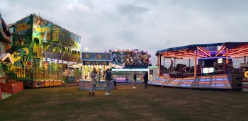 A lively amusement park scene features brightly lit rides and attractions. A fun house with vibrant lights and colorful murals stands to the left, while a spinning ride with red, blue, and purple lights is on the right. In the center, a booth offers games and prizes. People are casually walking and interacting, creating a festive atmosphere under a cloudy sky.