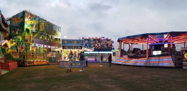 A lively amusement park scene features brightly lit rides and attractions. A fun house with vibrant lights and colorful murals stands to the left, while a spinning ride with red, blue, and purple lights is on the right. In the center, a booth offers games and prizes. People are casually walking and interacting, creating a festive atmosphere under a cloudy sky.