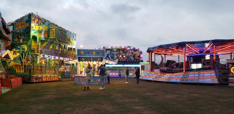 A lively amusement park scene features brightly lit rides and attractions. A fun house with vibrant lights and colorful murals stands to the left, while a spinning ride with red, blue, and purple lights is on the right. In the center, a booth offers games and prizes. People are casually walking and interacting, creating a festive atmosphere under a cloudy sky.