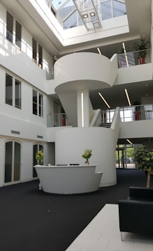A spacious and modern lobby area with a minimalist design. The focal point is a sleek, curved reception desk adorned with floral arrangements. The lobby has a large central staircase with a spiral design, leading to an upper level that features glass railings and large windows. The space is well-lit with natural light streaming in through a skylight, complemented by linear ceiling lights. The interior is decorated with potted plants and has a combination of white and dark flooring.